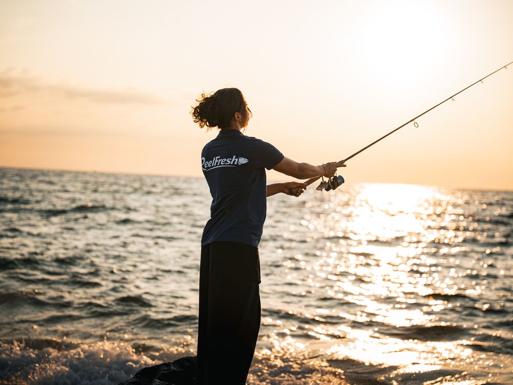 A woman casting a long rod out over the water with the low sun behind her