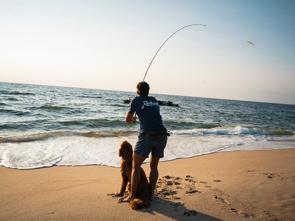 A fisherman mid-cast at the surf, rod bent, a dog sitting at his feet watching the line go out