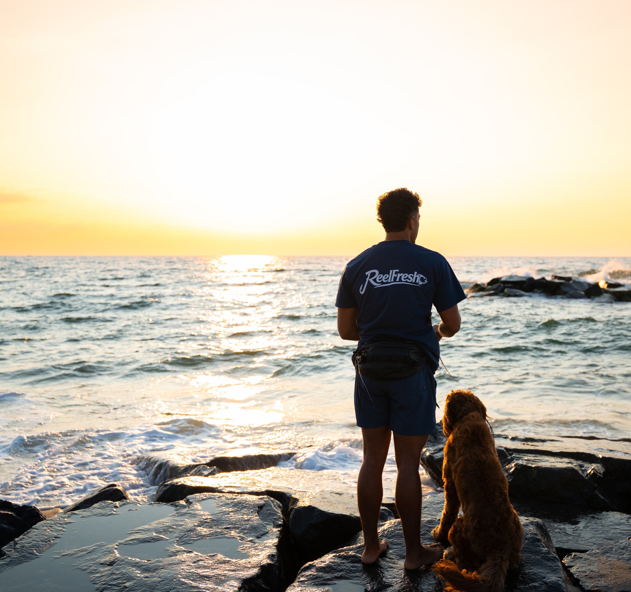 A fisherman in a ReelFresh shirt and a dog standing together on the jetty rocks, looking out at the water in golden light