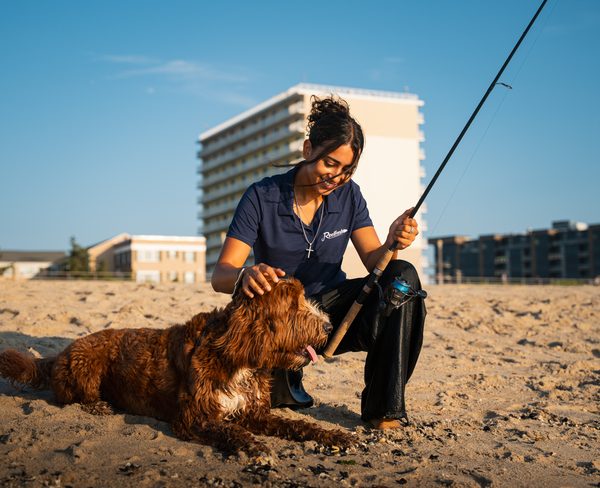 A woman in a ReelFresh polo kneeling on the sand, smiling and petting a wet, curly-haired dog, a rod in her other hand