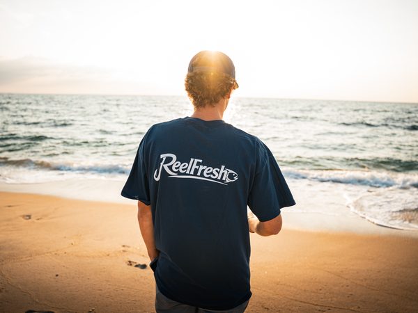 The ReelFresh wordmark across the back of a navy shirt, the wearer facing the surf