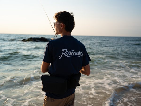 A fisherman with a sling pack and rod standing at the shoreline, looking down the beach