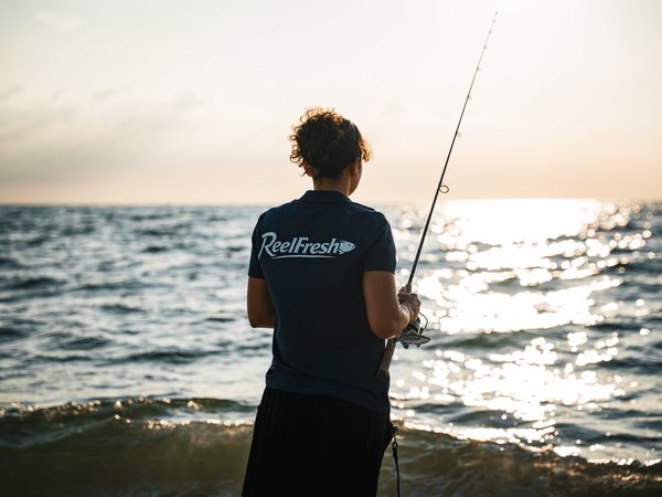 A woman with a rod facing the water, the surface sparkling in the low light