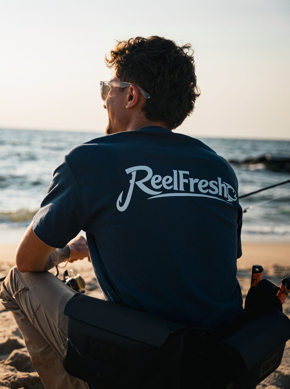 A fisherman in a ReelFresh tee sitting on the sand with his rod, looking out at the water at golden hour