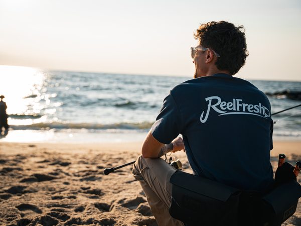 A fisherman sitting on the sand with his rod across his knees, looking out over the water