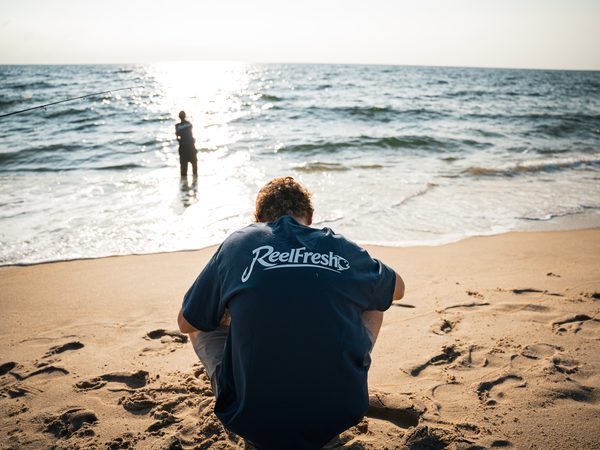 A fisherman sitting on the beach while another stands in the shallows, the sun bright on the water