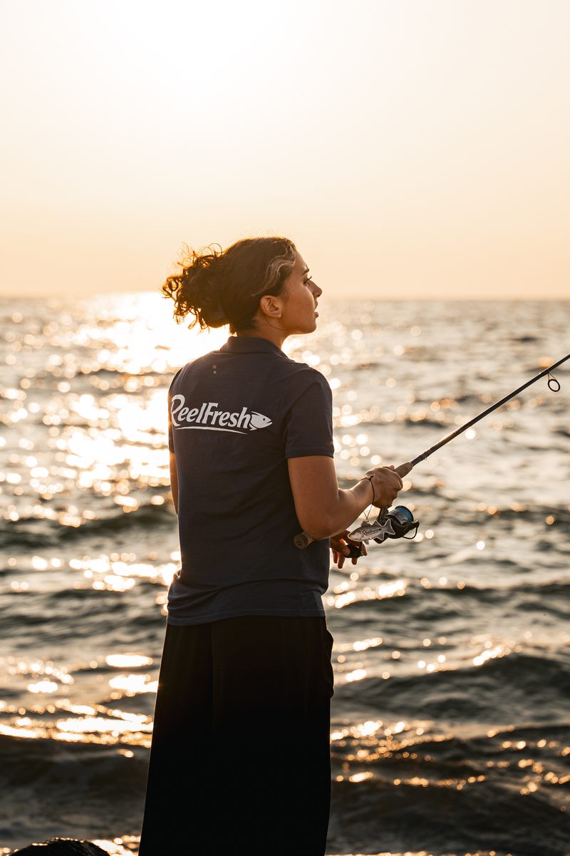 A woman in a ReelFresh polo holding a rod at the water's edge, the freshener hanging from the reel