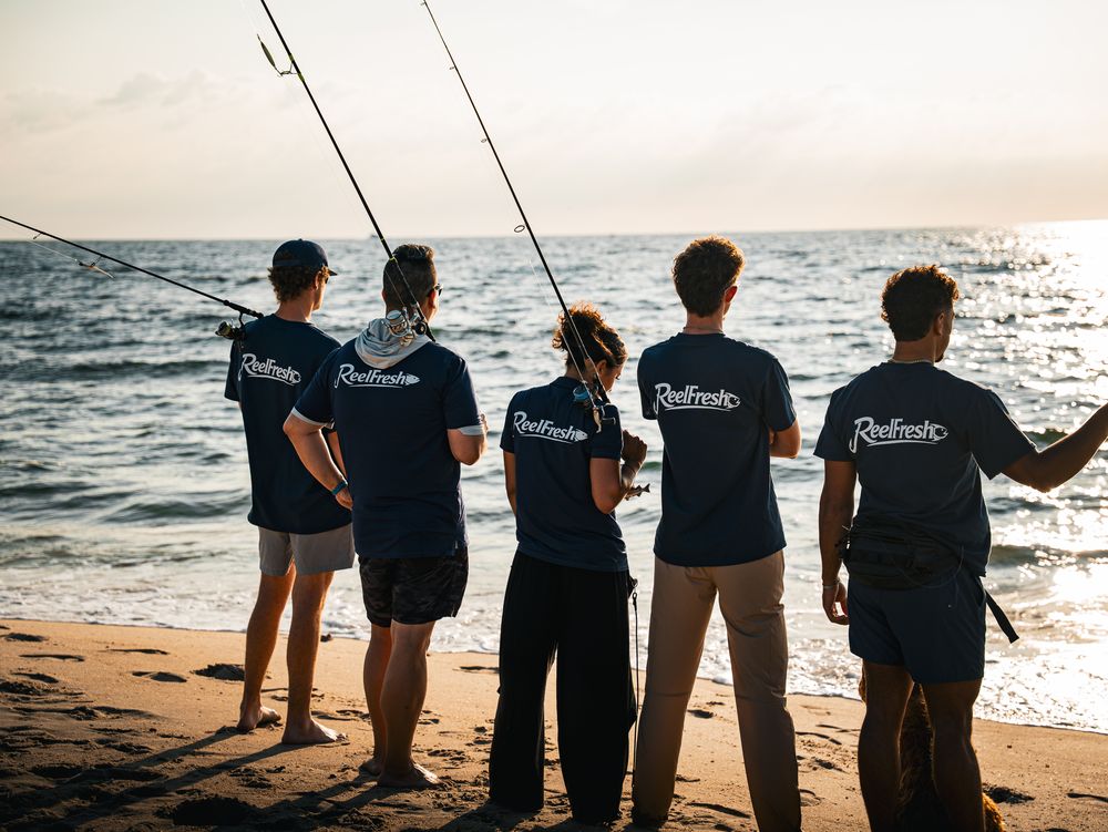 Five fishermen with rods over their shoulders, lined up at the shoreline looking out to sea