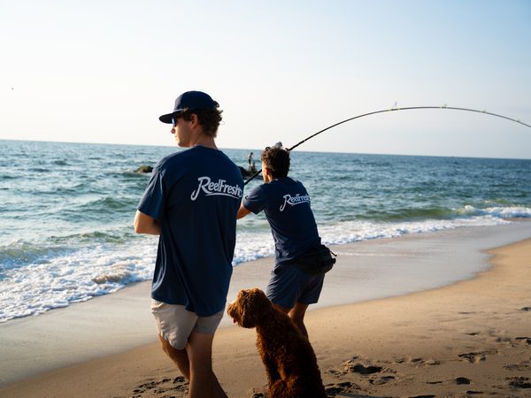 Two fishermen at the shoreline, one casting with a bent rod, a dog sitting between them