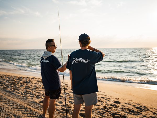 Two friends in ReelFresh shirts talking on the beach, one holding a surf rod upright between them