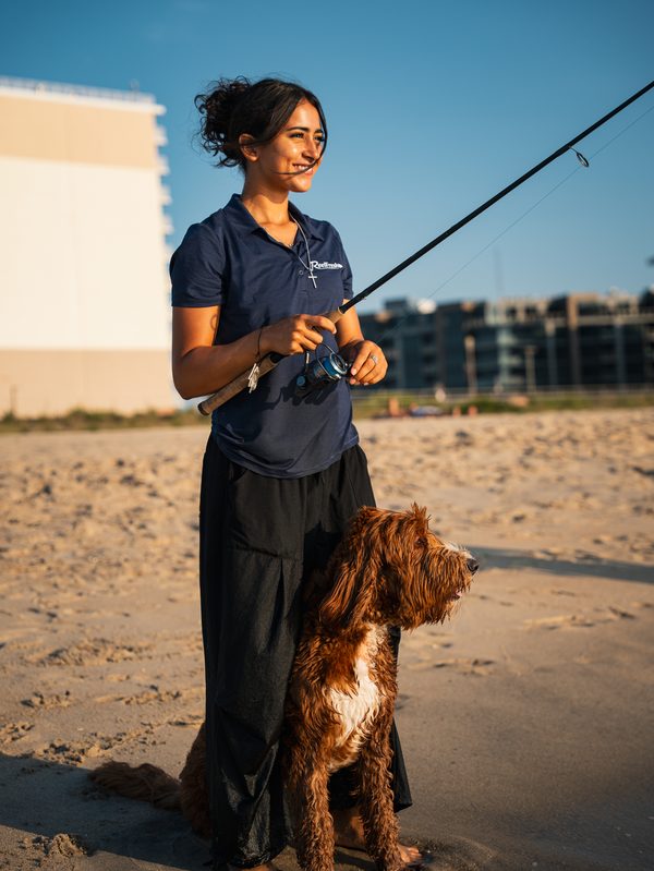 A woman standing on the beach with a rod and reel, a dog sitting at her side, both looking toward the water